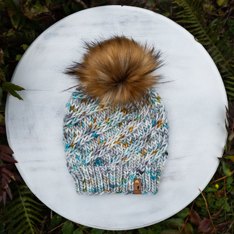 Knitted beanie with a pom-pom on a white surface surrounded by green foliage