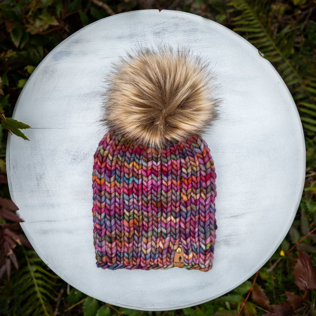 Knitted beanie with a pom-pom on a white surface surrounded by green foliage