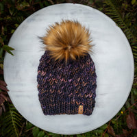 Knitted beanie with a large pom-pom on a white surface surrounded by green foliage