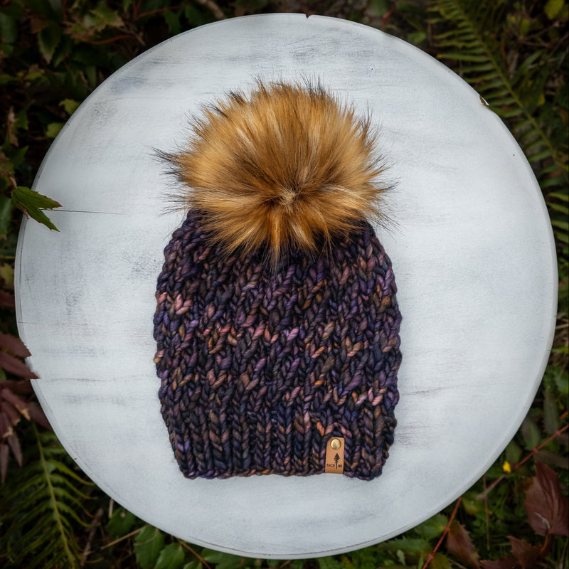 Knitted beanie with a large pom-pom on a white surface surrounded by green foliage