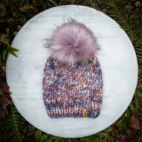 Knitted hat with a pink pom-pom on a white surface surrounded by green foliage