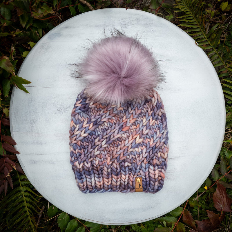 Knitted hat with a pink pom-pom on a white surface surrounded by green foliage