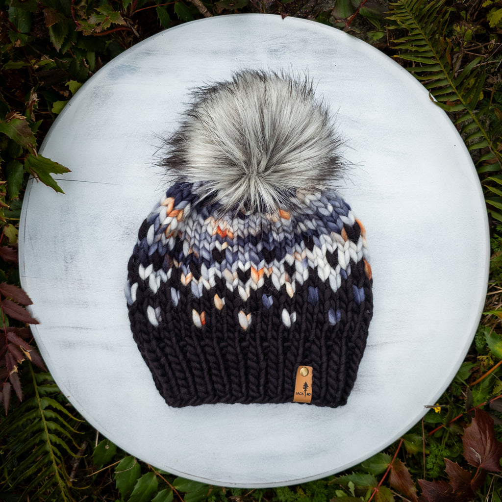 Knitted beanie with a pom-pom on a white surface surrounded by green foliage