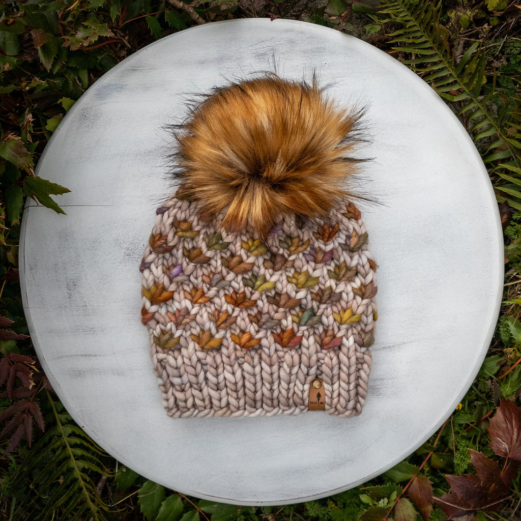 Knitted beanie with lotus flower pattern and fluffy Caramel faux fur pom-pom on a white surface.