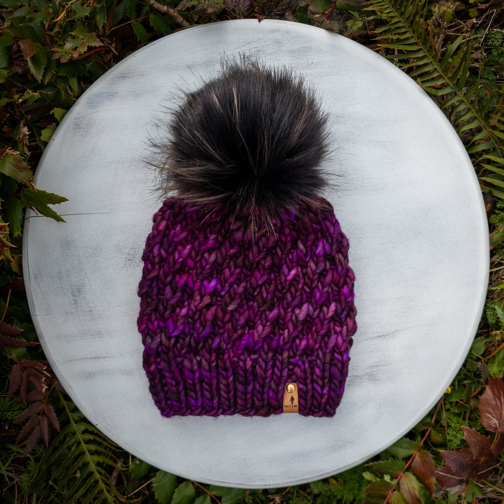 Purple knit toque with a large black pom-pom on a white surface surrounded by green foliage.
