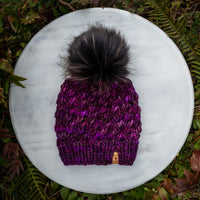 Purple knit toque with a large black pom-pom on a white surface surrounded by green foliage.
