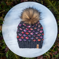 Knitted grey beanie with colorful lotus flower pattern and faux fur pom-pom on a white surface