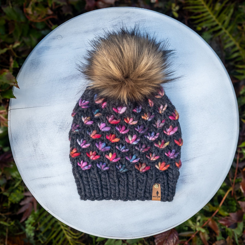 Knitted grey beanie with colorful lotus flower pattern and faux fur pom-pom on a white surface