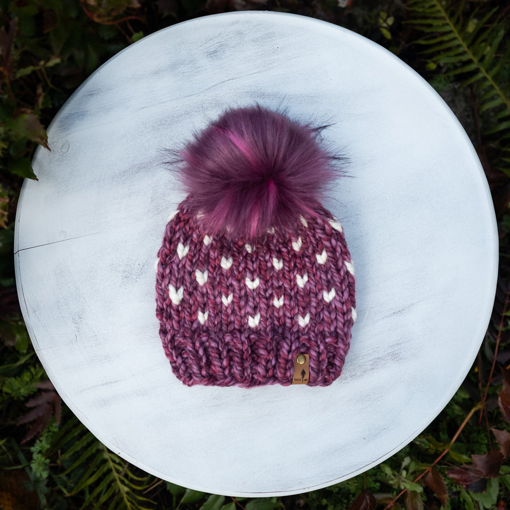 Purple knit beanie with a pink pom-pom on a white surface surrounded by green foliage.