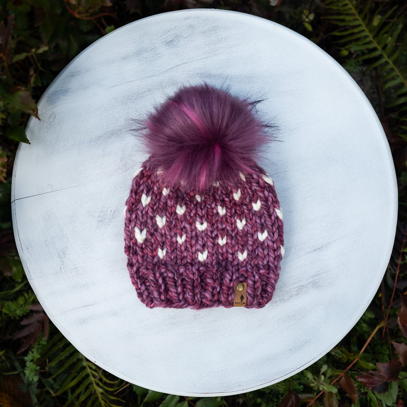 Purple knit beanie with a pink pom-pom on a white surface surrounded by green foliage.