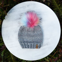 Gray knit beanie with a colorful pom-pom on a white surface surrounded by green foliage.