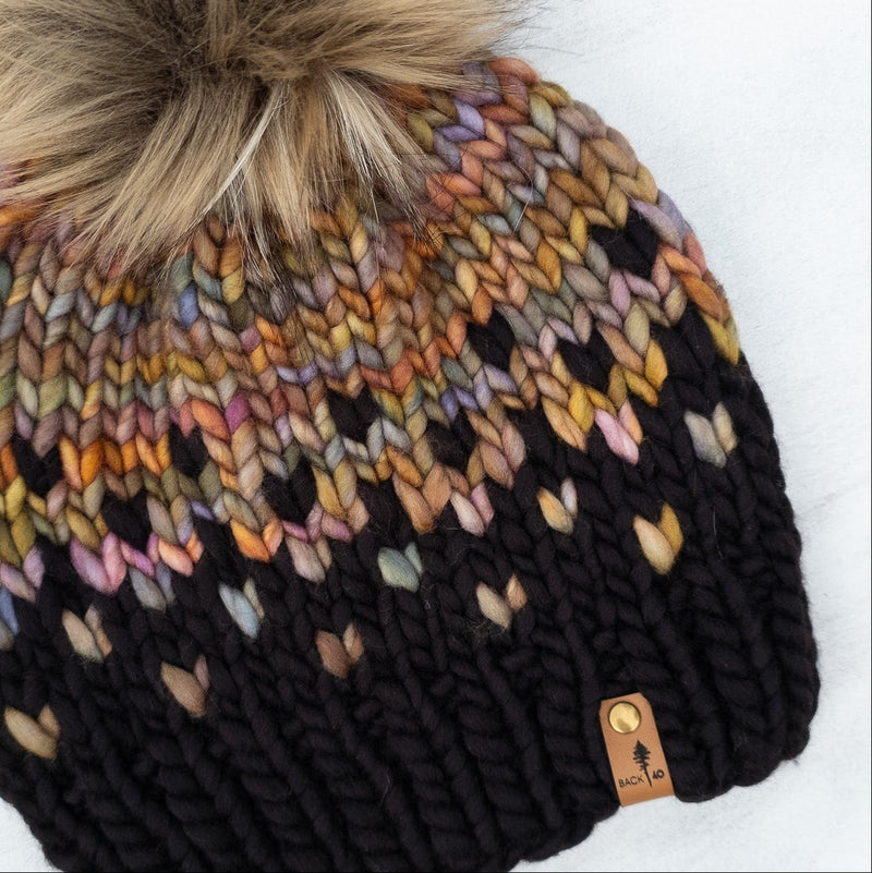 Knitted beanie with a colorful pattern and fur pom-pom on a white surface.