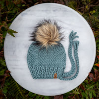 Knitted teal hat with a fluffy pom-pom on a white surface surrounded by greenery.