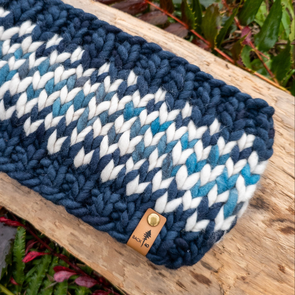 Knitted headband with blue and white pattern on a wooden log with green foliage in the background