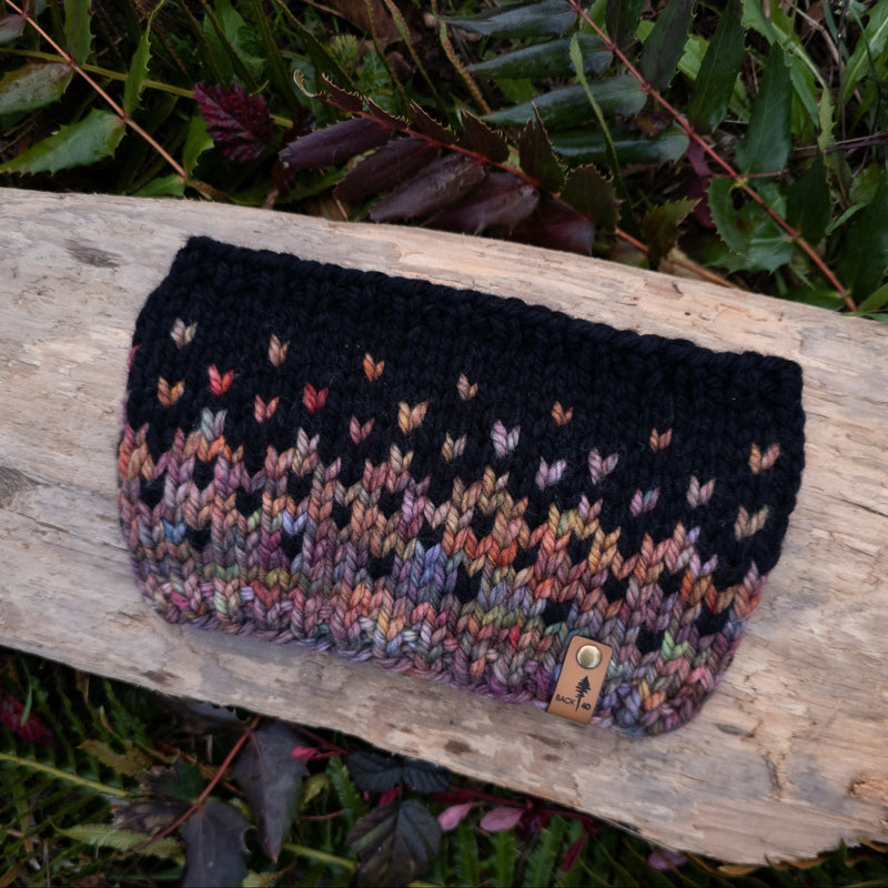 Knitted pouch with multicolor pattern on a wooden log with green foliage in the background