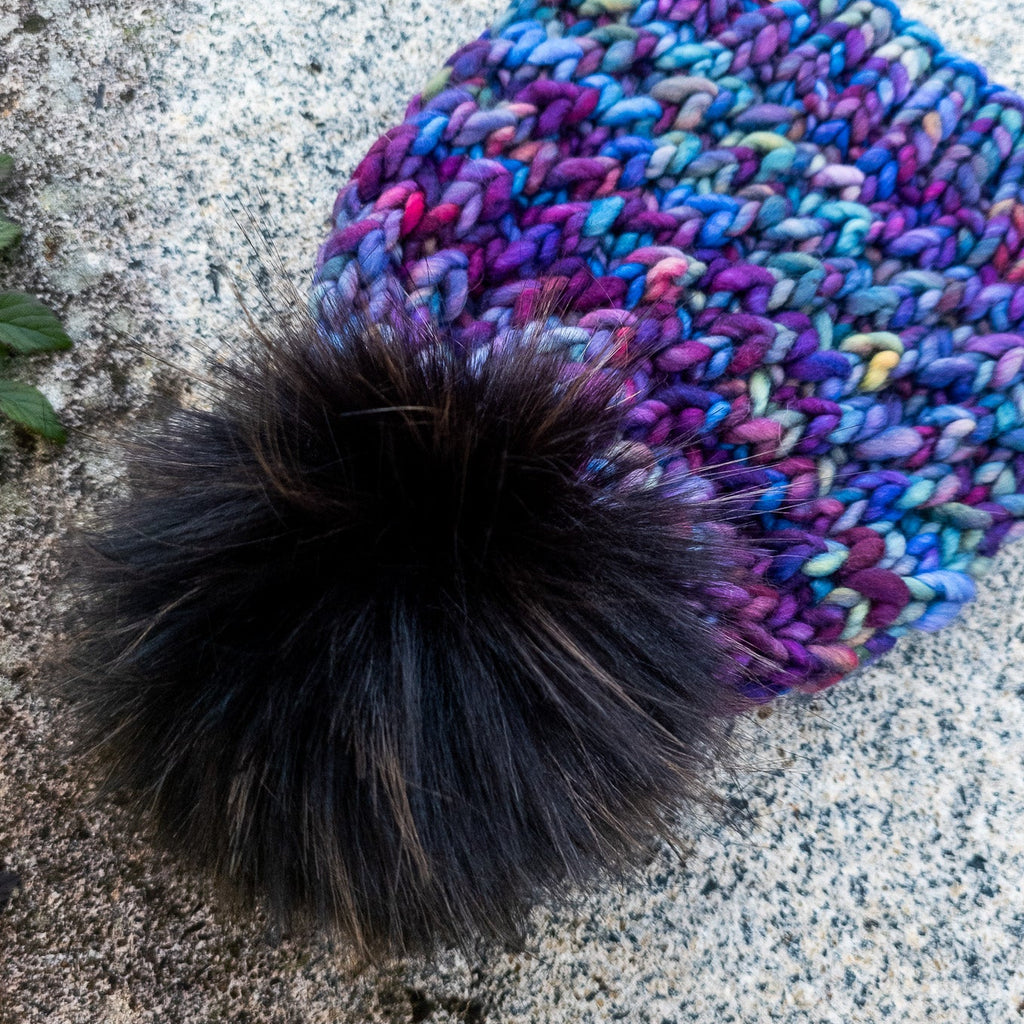 Multicolored knit beanie with a large black pom-pom on a stone surface.