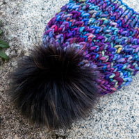 Multicolored knit beanie with a large black pom-pom on a stone surface.